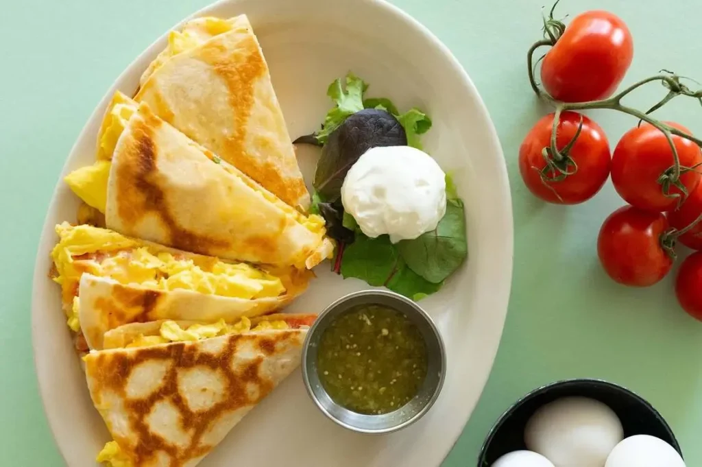 A golden-brown breakfast quesadilla (kw) filled with scrambled eggs, melted cheese, and colorful veggies, sliced and served with salsa on a plate